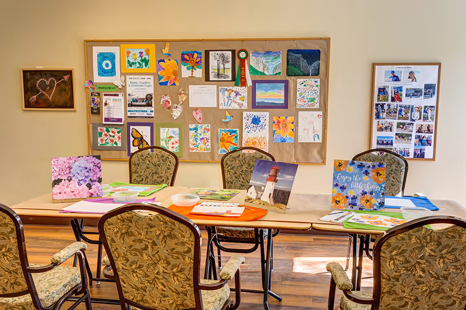 A room with a rectangular table surrounded by six floral-patterned chairs. On the table are various colorful art supplies and paintings, including images of flowers and a lighthouse. Behind the table is a bulletin board displaying multiple pieces of artwork, drawings, and notices. The room has wooden flooring and light-colored walls.