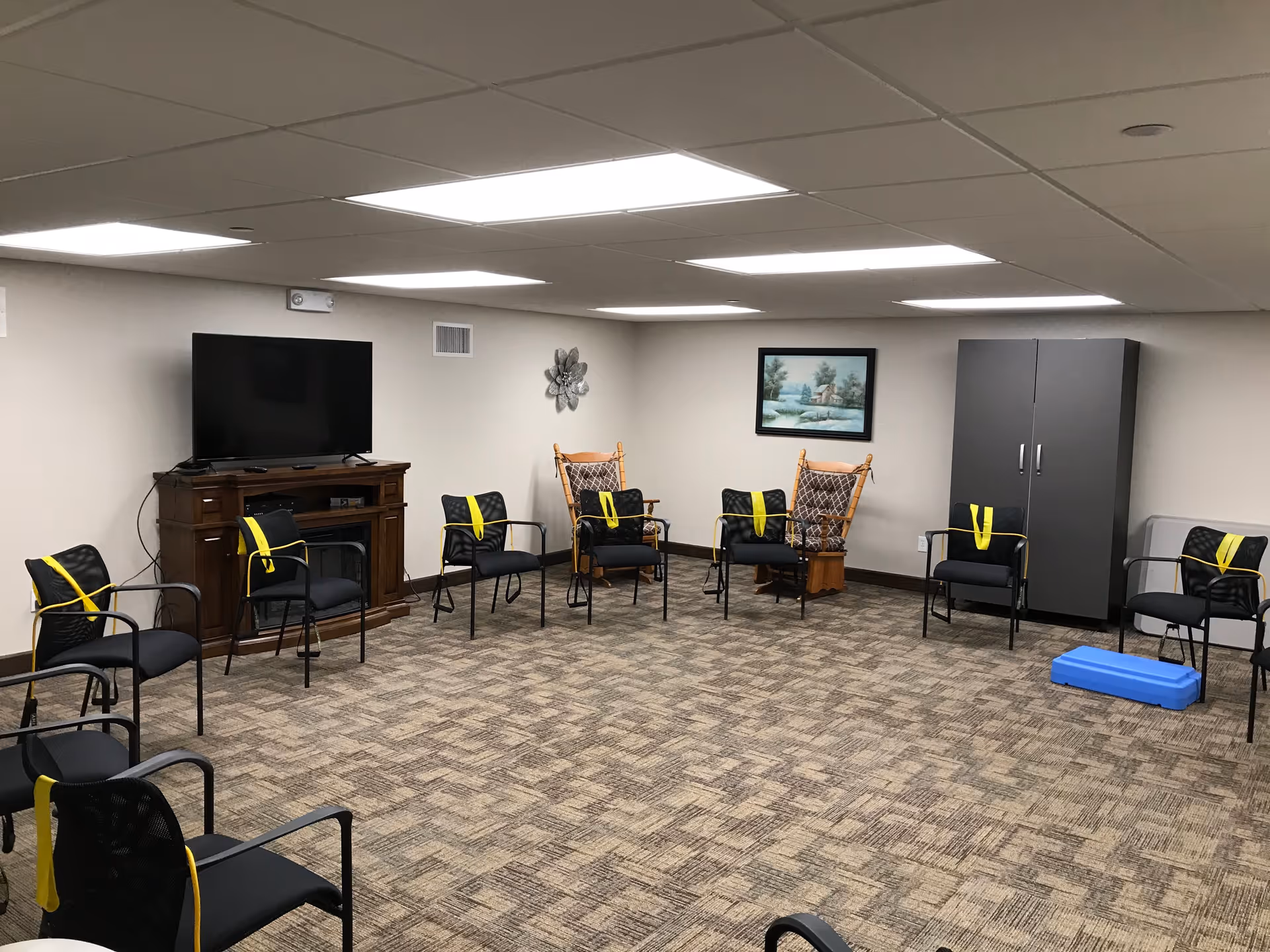 A room with chairs arranged in a circle, each chair has a yellow resistance band draped over it. The room has a carpeted floor, a flat-screen TV on a wooden stand, two wooden rocking chairs, a gray cabinet, a blue step platform, and a framed landscape painting on the wall.