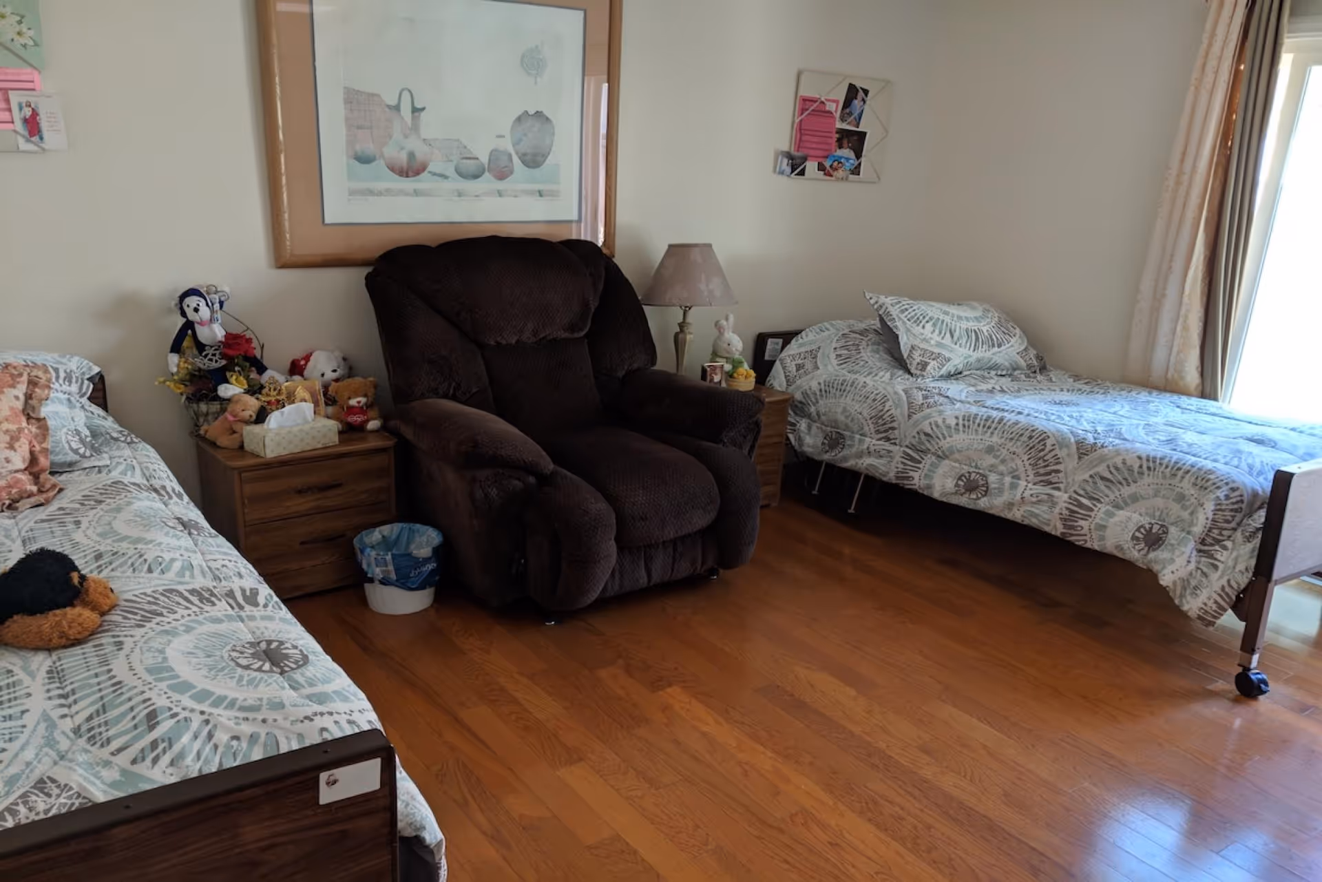 A cozy bedroom with two single beds on either side of a dark brown recliner chair. Each bed has patterned bedding in shades of blue and gray. Between the beds are wooden nightstands, one with a lamp and decorative items, and the other with stuffed animals and a tissue box. The room has wooden flooring and light-colored walls with framed pictures and a window with curtains letting in natural light.