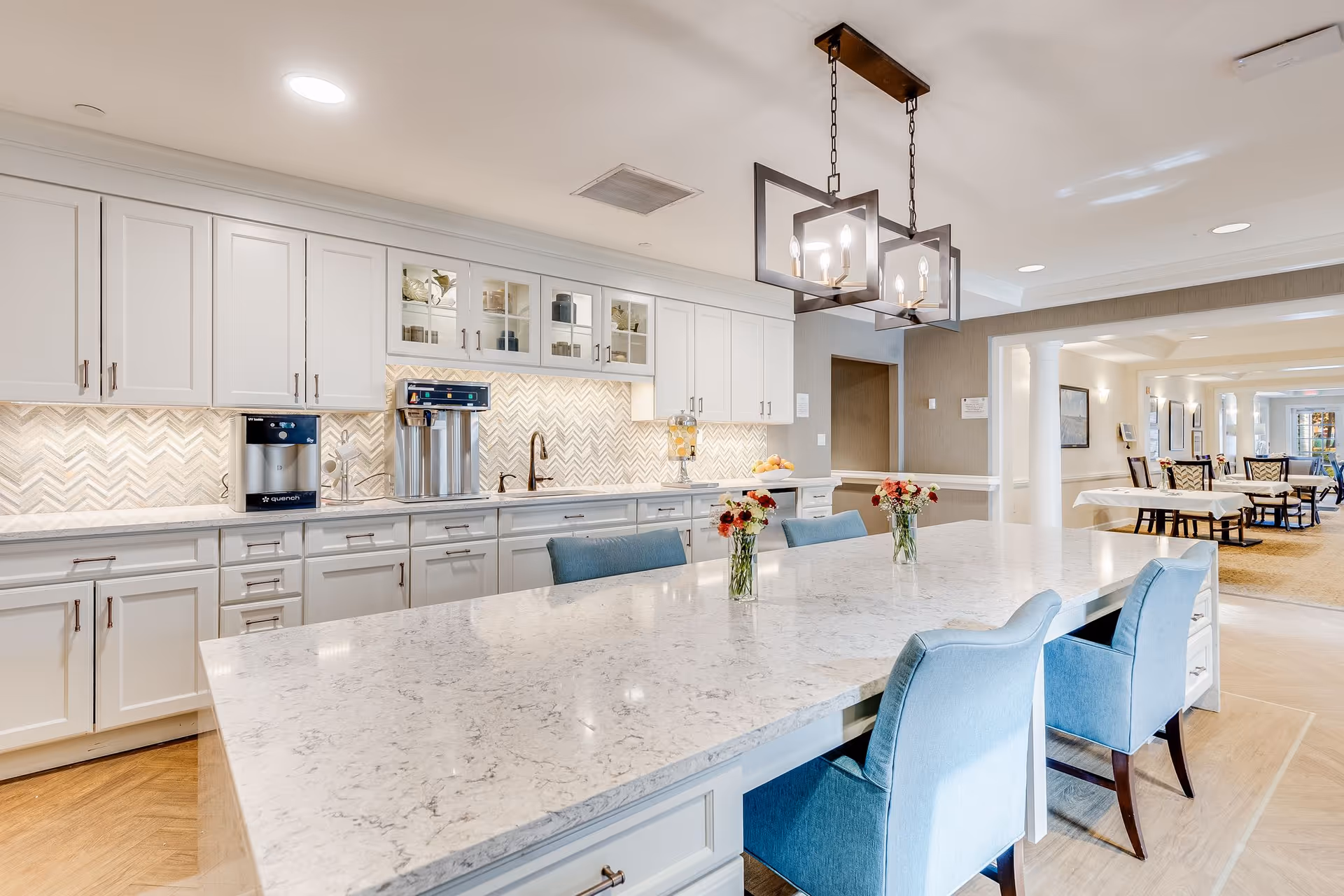 Bright communal kitchen with a large marble island, blue upholstered chairs, white cabinets and pendant lighting.
