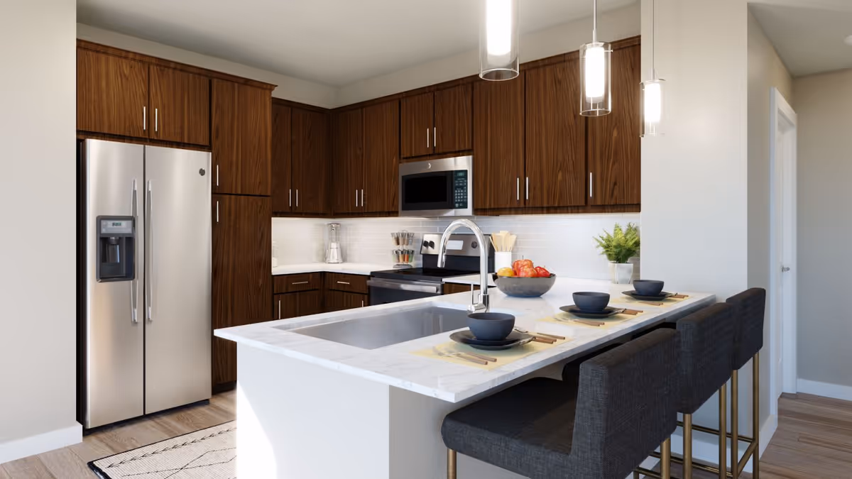 Modern kitchen with dark wood cabinets, stainless steel refrigerator, microwave, and stove. A white marble countertop island with a built-in sink and three dark upholstered bar stools is set with black bowls, plates, and chopsticks. Pendant lights hang above the island, and a bowl of fruit and a small potted plant are on the counter.