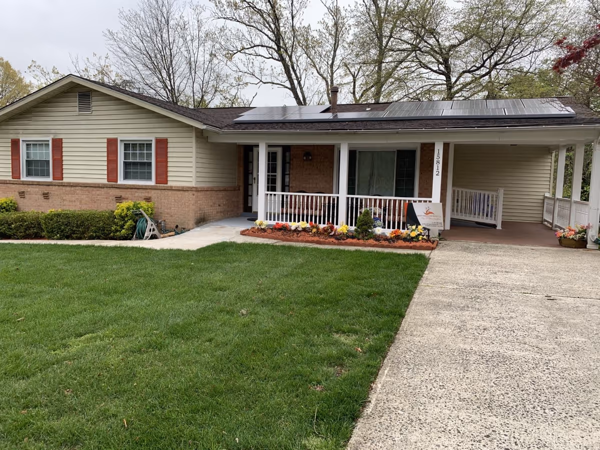 Single-story house with beige siding and brick lower walls, featuring a front porch with white railings and a flower bed with colorful flowers. Solar panels are installed on the roof. The house number 15812 is visible on a porch column. A concrete driveway leads up to the porch, and there is a green lawn in front of the house with some bushes along the side.