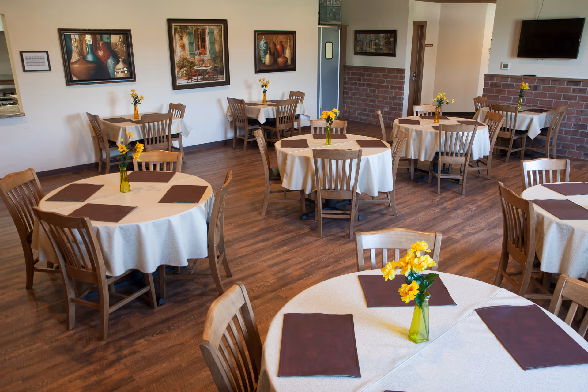 Dining room with round tables covered in tablecloths, wooden chairs, and yellow flower centerpieces.