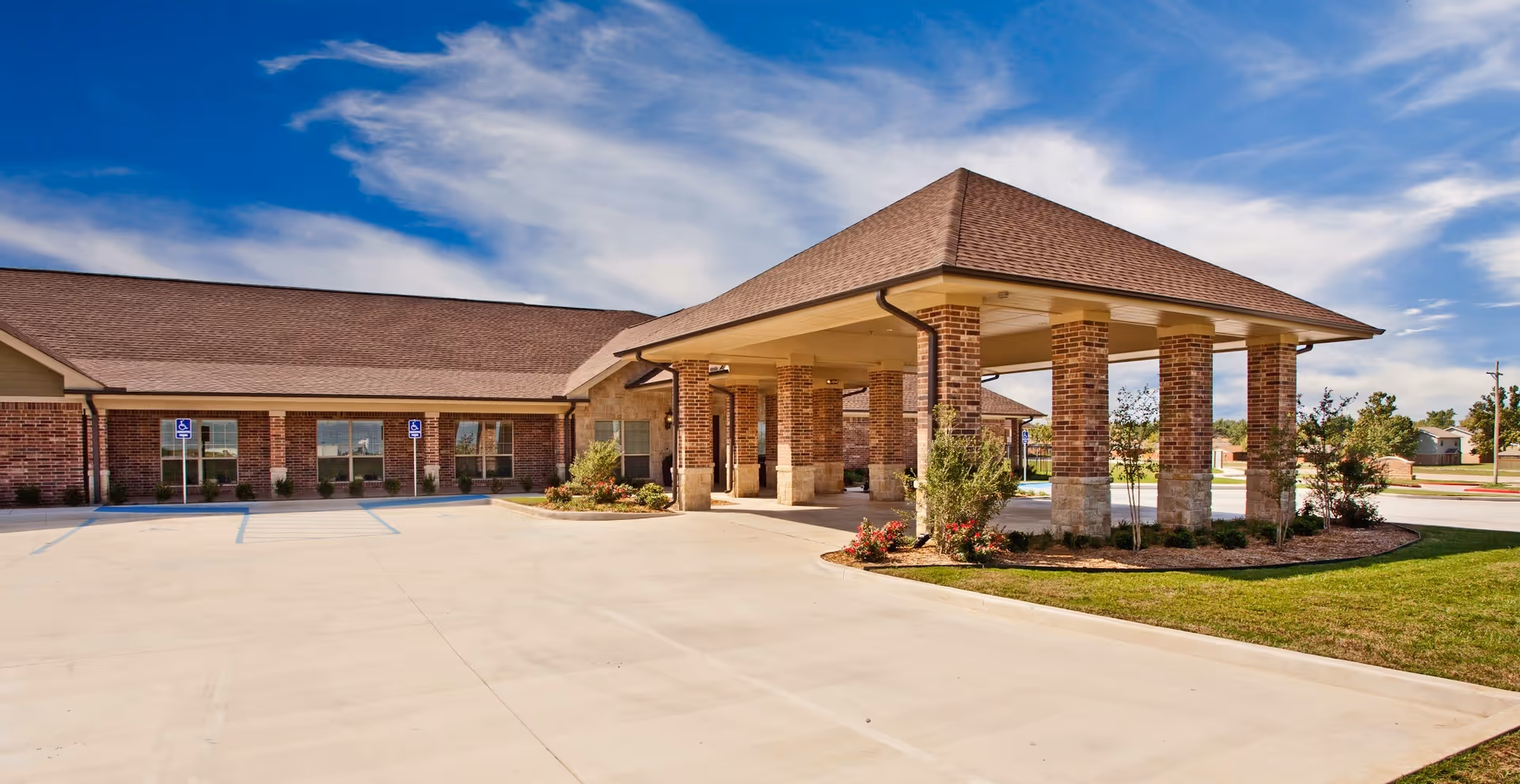 Front exterior of a single-story brick rehabilitation and skilled care building with a covered entrance/porte-cochere, parking area and landscaped grounds under a blue sky.