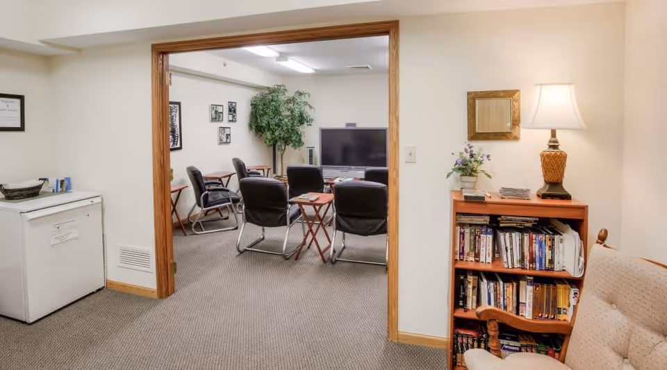 Interior view of a senior living facility showing a small room with a white freezer on the left and a doorway leading to a lounge area with black chairs arranged around a TV. On the right, there is a wooden bookshelf filled with books and a table lamp on top, next to a cushioned wooden chair.