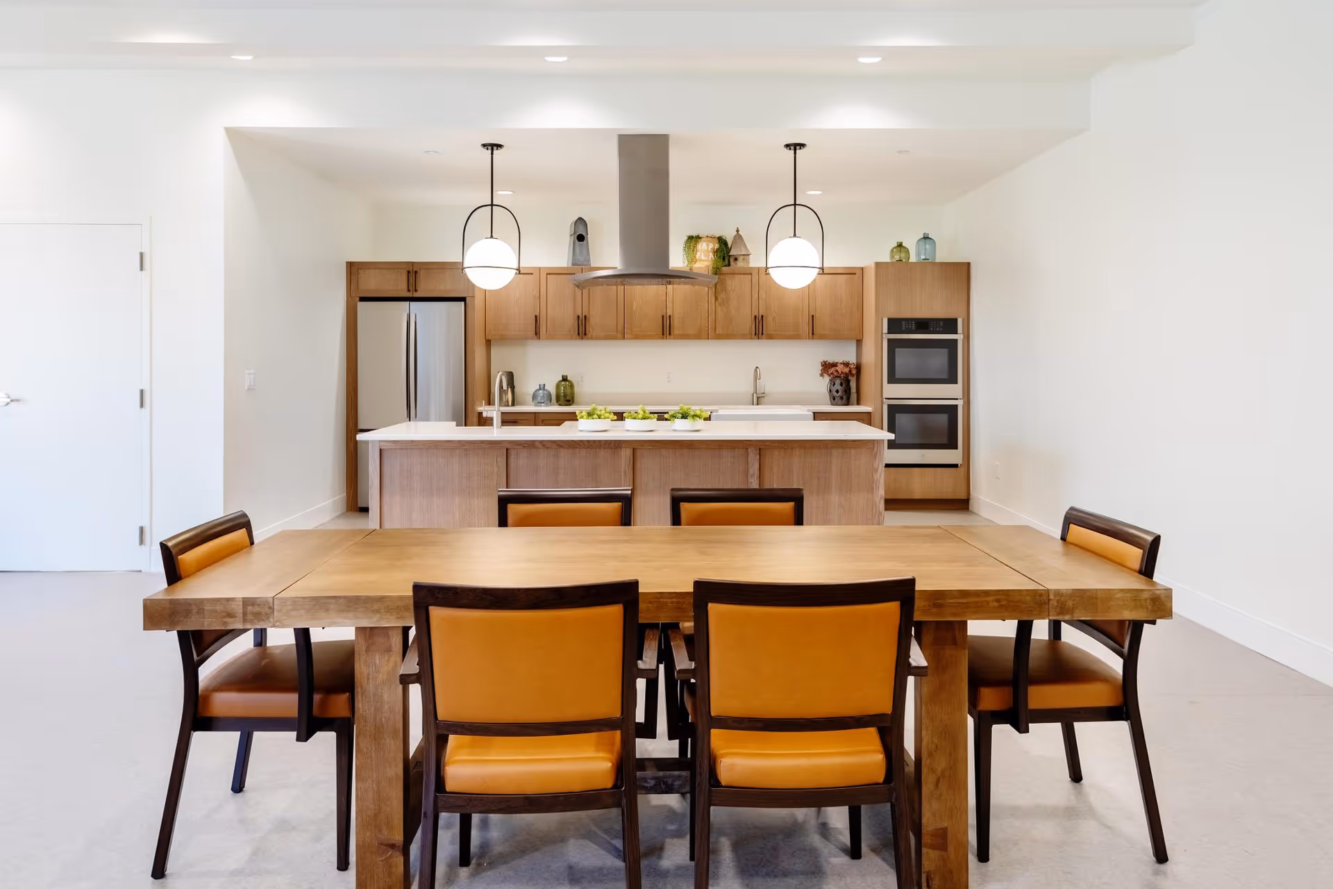 Modern communal dining area with a wooden table and chairs facing a kitchen island with pendant lights and stainless steel appliances.