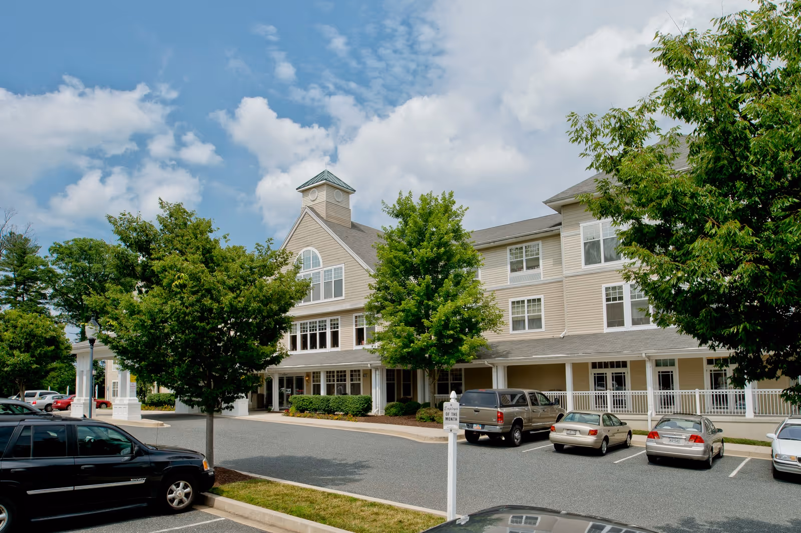 Exterior view of a multi-story senior living facility building with beige siding, large windows, a small tower feature, and a covered entrance. Several cars are parked in the parking lot in front of the building, and there are green trees and shrubs around the area under a partly cloudy sky.