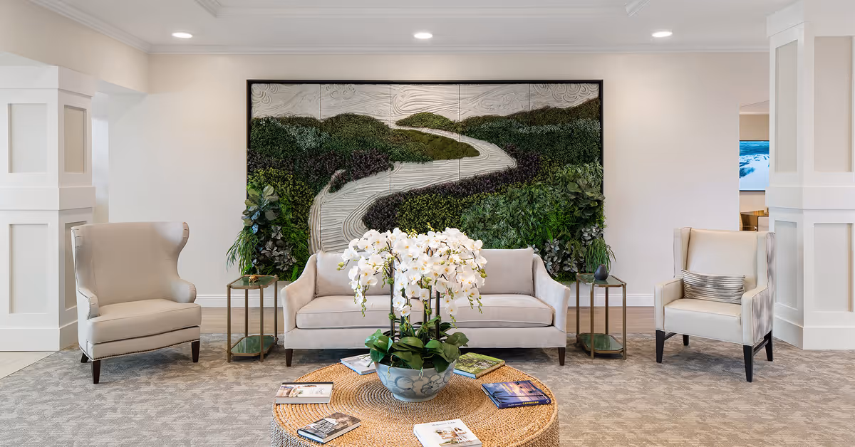 Bright communal seating area with a sofa flanked by two armchairs, a round wicker coffee table topped with books and a potted orchid, and a large nature-themed wall art behind.