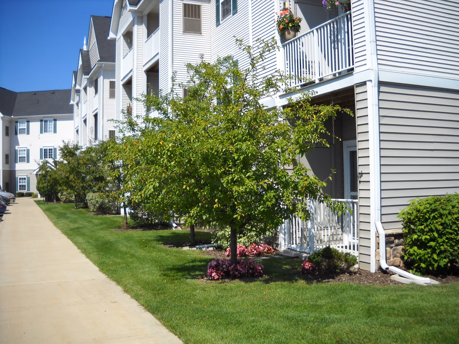 Exterior view of a multi-story residential building with white siding and balconies. There is a well-maintained lawn with small trees and flower beds along a concrete walkway. The sky is clear and blue.