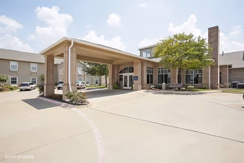 Exterior view of Morada Pantego facility showing the main entrance with a covered drop-off area, brick building facade, large windows, a tree, and a clear sky with some clouds.