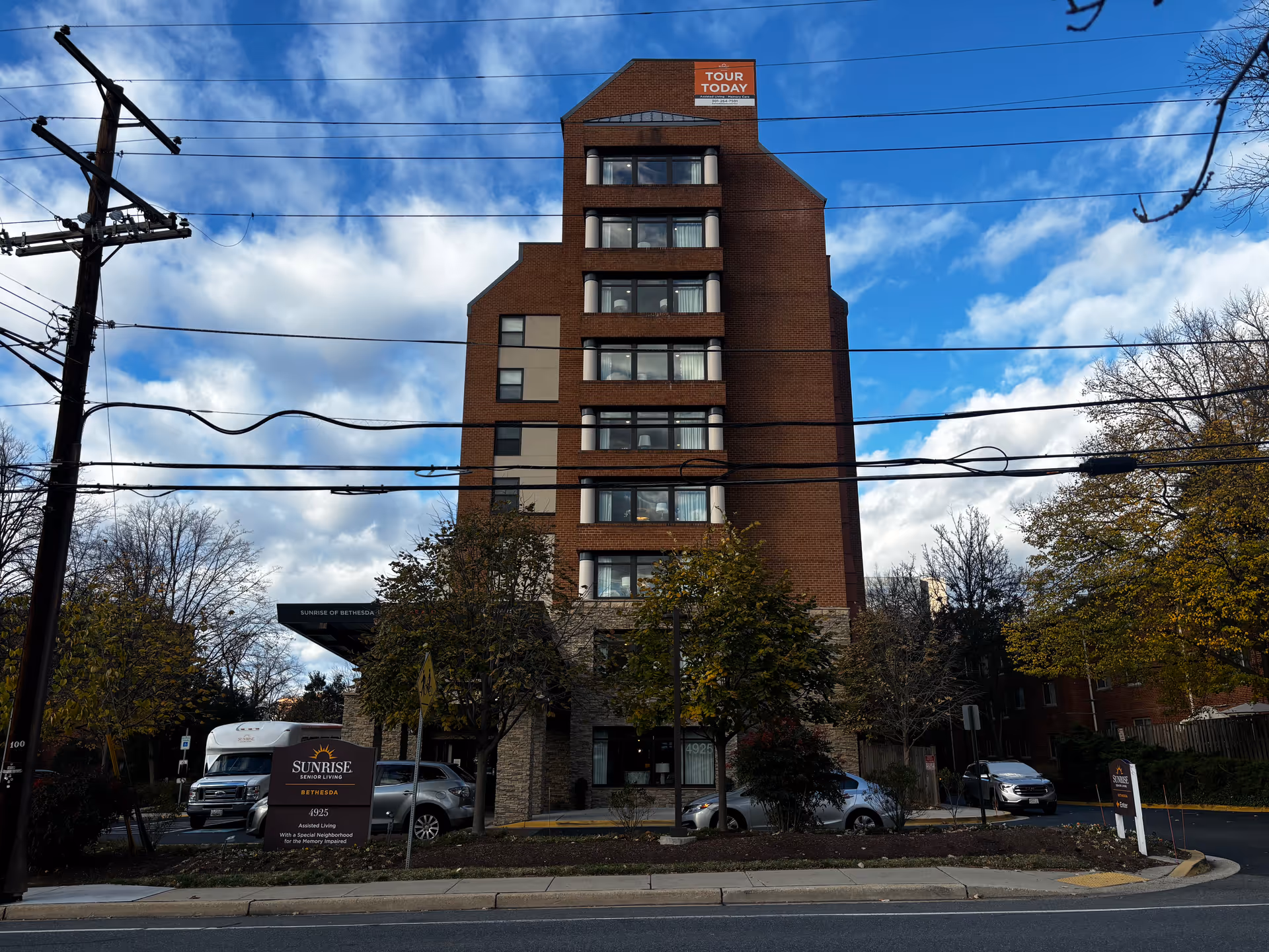 Exterior view of a multi-story brick building with a sign in front that reads 'Sunrise Senior Living Bethesda, 4925, Assisted Living with a Special Neighborhood for the Memory Impaired.' The building has several windows and a sign at the top that says 'Tour Today.' Trees and parked cars are visible around the building under a partly cloudy sky.