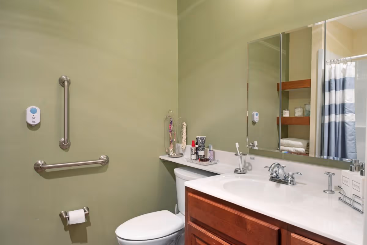 A bathroom with light green walls featuring a white toilet and a wooden vanity with a white countertop and sink. There are two stainless steel grab bars on the wall, a toilet paper holder, and a wall-mounted emergency call button. On the countertop, there are various toiletries and a jewelry stand. A large mirror is above the sink, reflecting shelves with towels and jars, and a shower curtain with blue and white stripes is visible in the background.