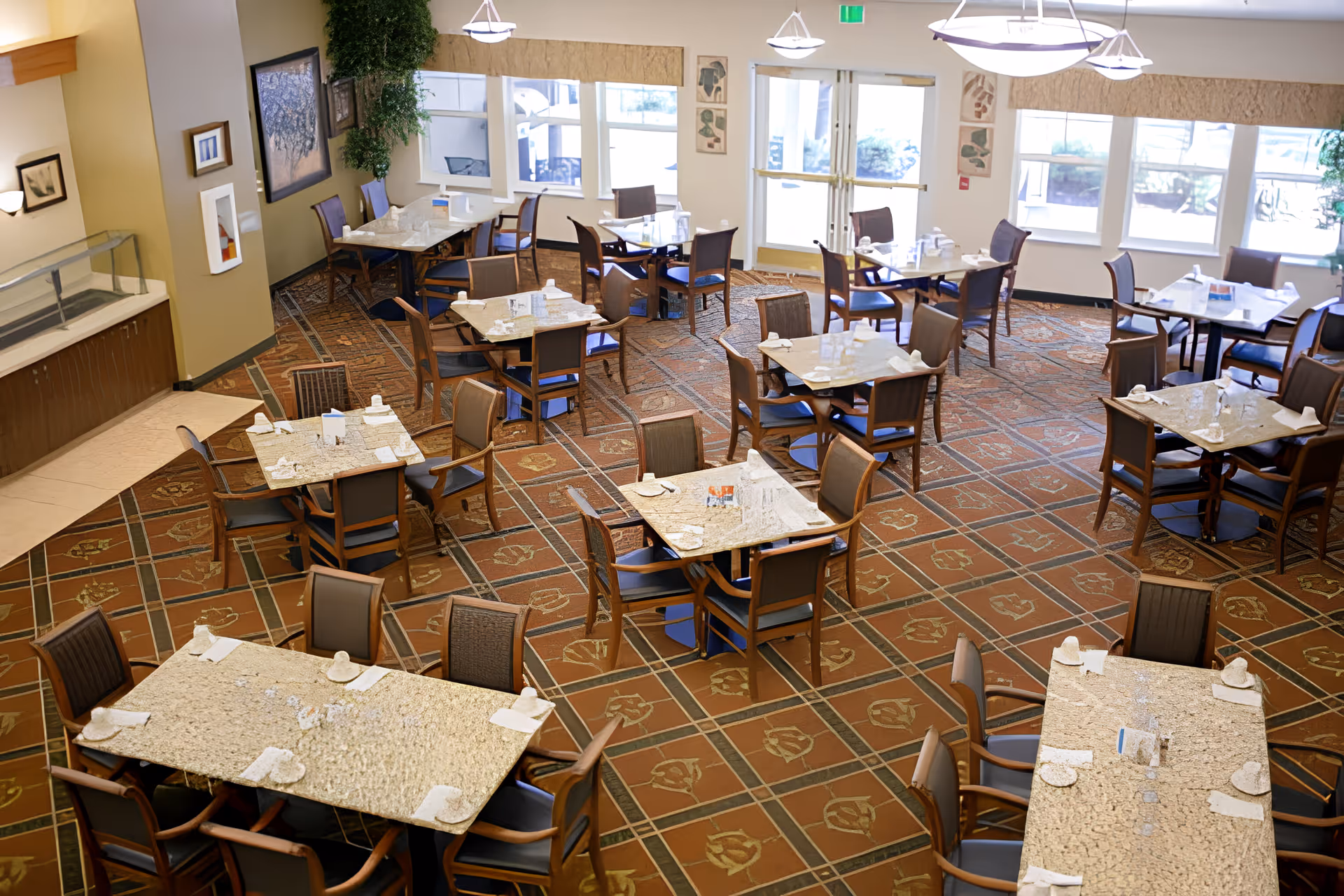 A spacious dining room in a senior living facility with multiple tables and chairs arranged neatly. Each table is set with napkins, plates, and glasses. Large windows allow natural light to fill the room, and there are decorative plants and framed artwork on the walls.