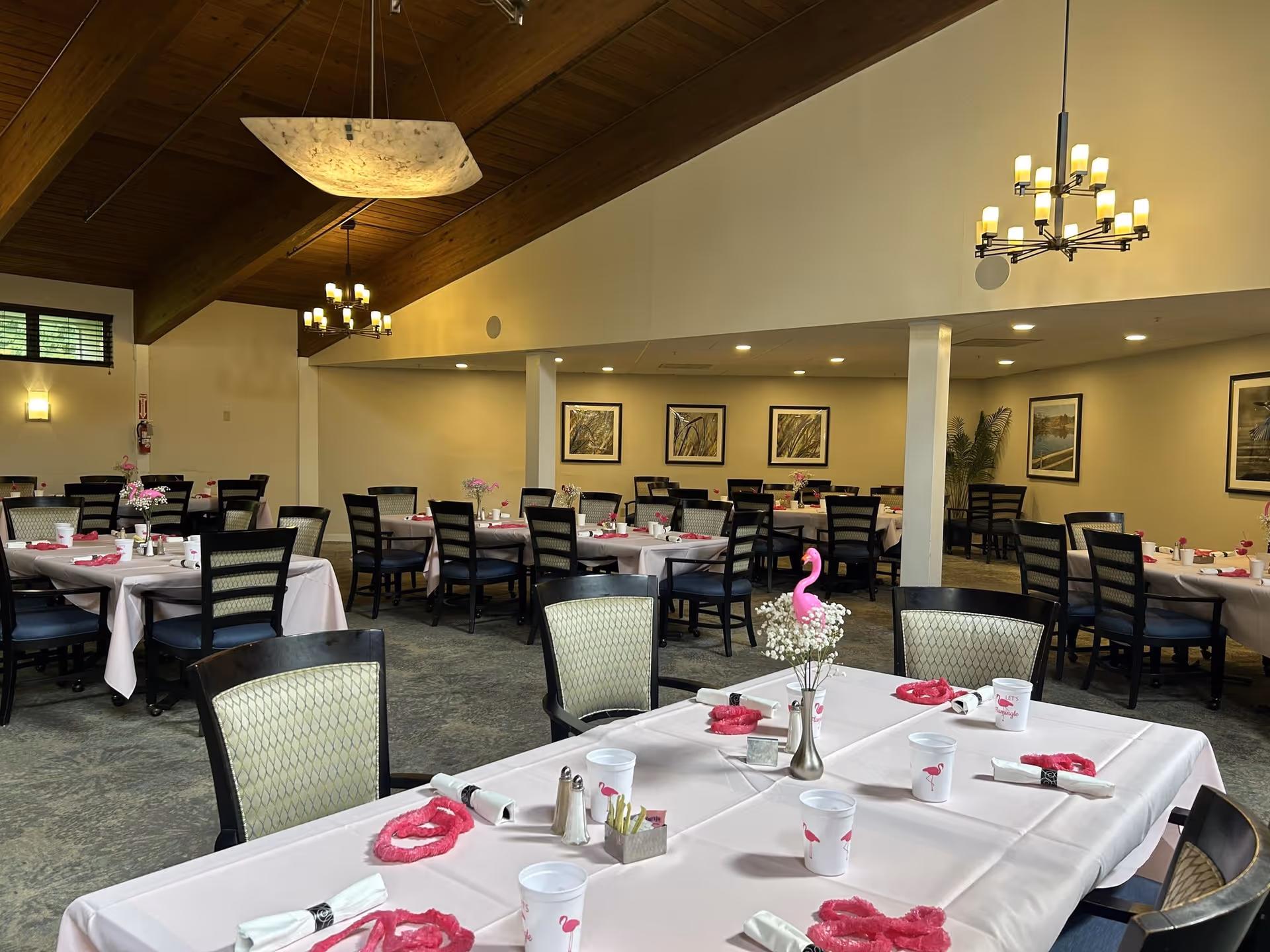 Dining room with tables dressed in pink decorations and place settings, chairs, and chandeliers beneath a vaulted wooden ceiling.