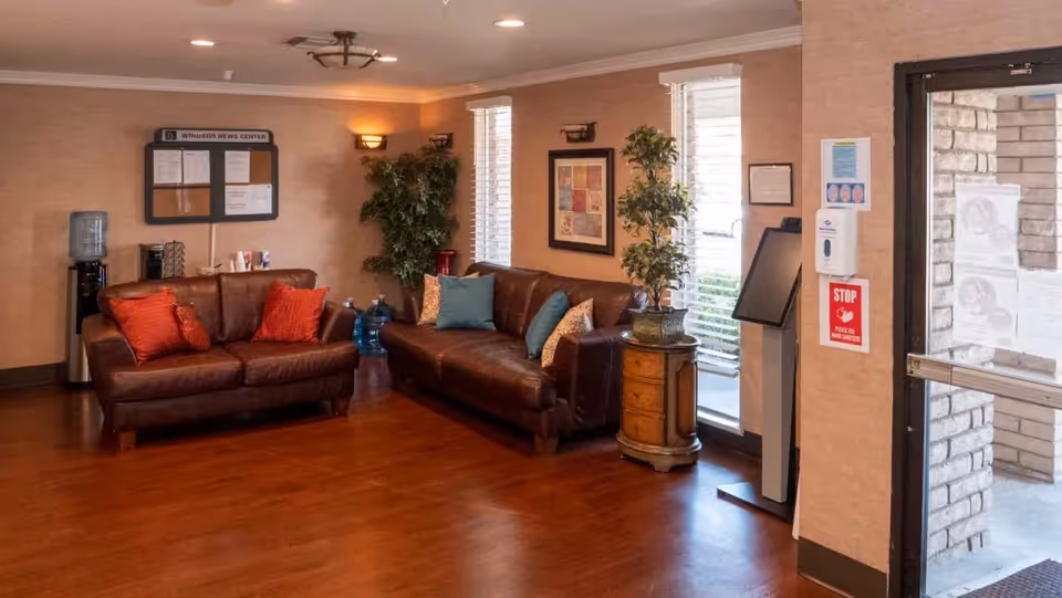 A cozy living room area with two brown leather sofas adorned with colorful cushions, a wooden side table with a potted plant, a water dispenser, and a bulletin board on the wall. The room has wooden flooring, beige walls, and large windows letting in natural light. There is also a hand sanitizer dispenser and a red stop sign near the entrance door.