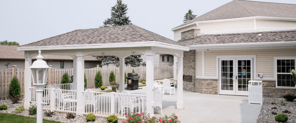 Outdoor patio area at Renaissance by Rennes - Marinette featuring a white gazebo with a shingled roof, white chairs and tables underneath, a grill, and a landscaped garden with flowers and shrubs. The building exterior has beige siding with stone accents and double glass doors.