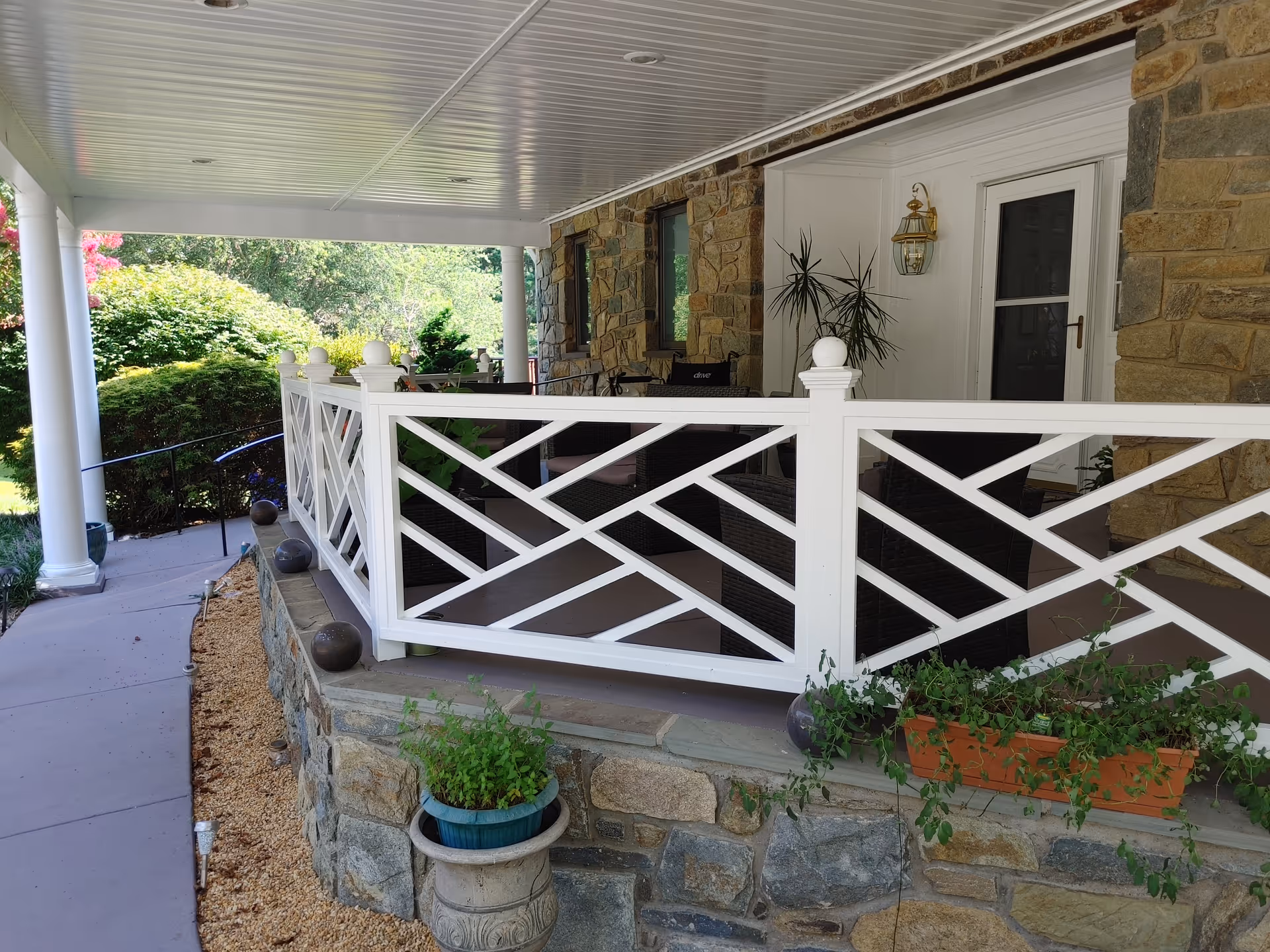 A covered porch area with white decorative railing and stone foundation. The porch has outdoor seating with dark wicker chairs and a table. There are potted plants on the porch and along the stone foundation. The porch is attached to a building with stone walls and a white door with a glass window. A pathway with a handrail leads up to the porch, surrounded by greenery and bushes.