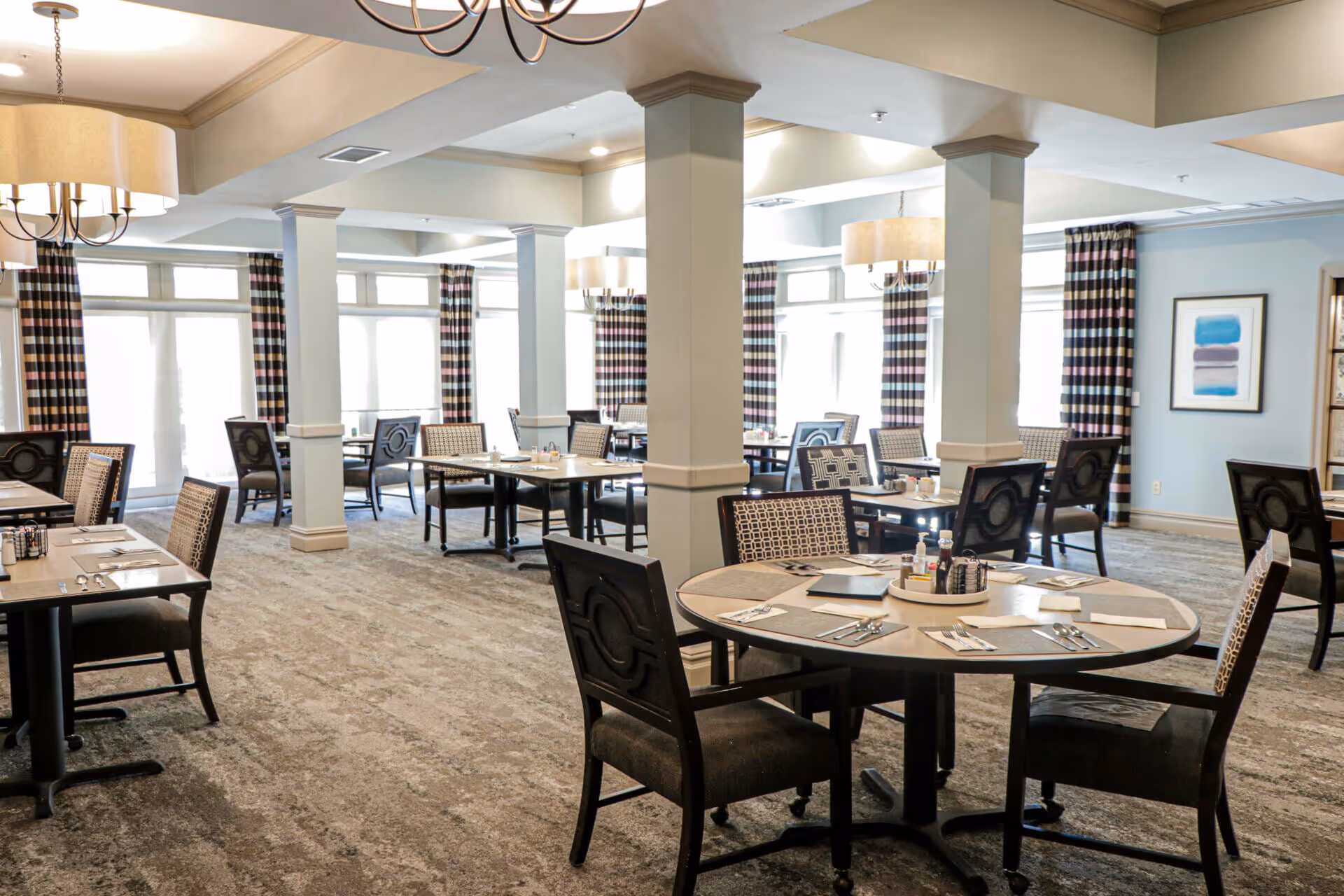 A spacious dining room with multiple tables and chairs arranged neatly. The room features large windows with striped curtains allowing natural light to fill the space. There are chandeliers hanging from the ceiling and the tables are set with placemats, utensils, and condiments.