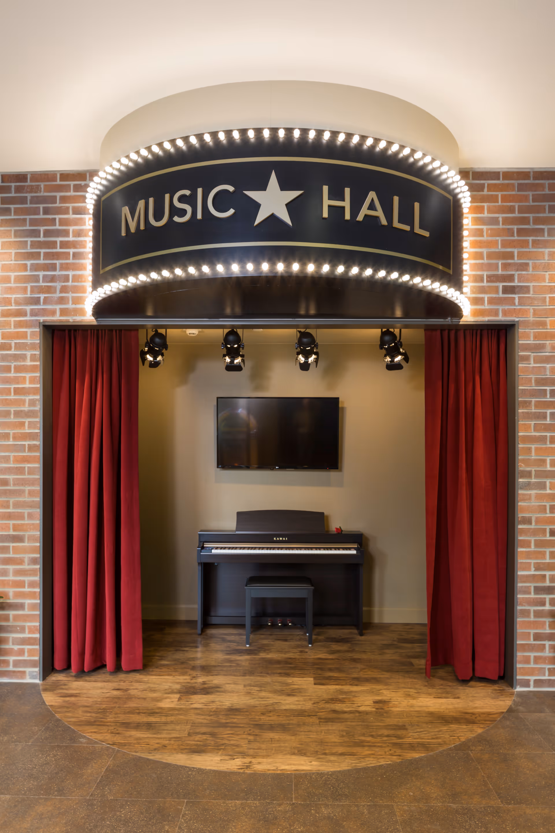 A small stage area labeled 'Music Hall' with a black upright piano and matching bench. The stage is framed by red curtains and overhead spotlights, with a flat screen TV mounted on the wall behind the piano. The floor is wood and the surrounding walls are brick.