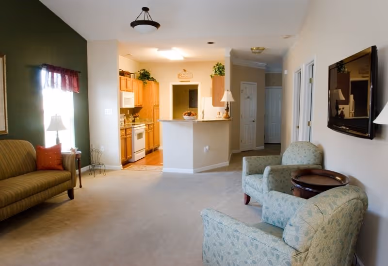 A cozy living room area with two patterned armchairs and a round wooden side table, a wall-mounted flat screen TV, and a striped sofa with a red pillow near a window with a red valance. The room opens into a kitchen with wooden cabinets, a white stove, microwave, and refrigerator. The walls are painted in neutral tones with one green accent wall, and the floor is carpeted.