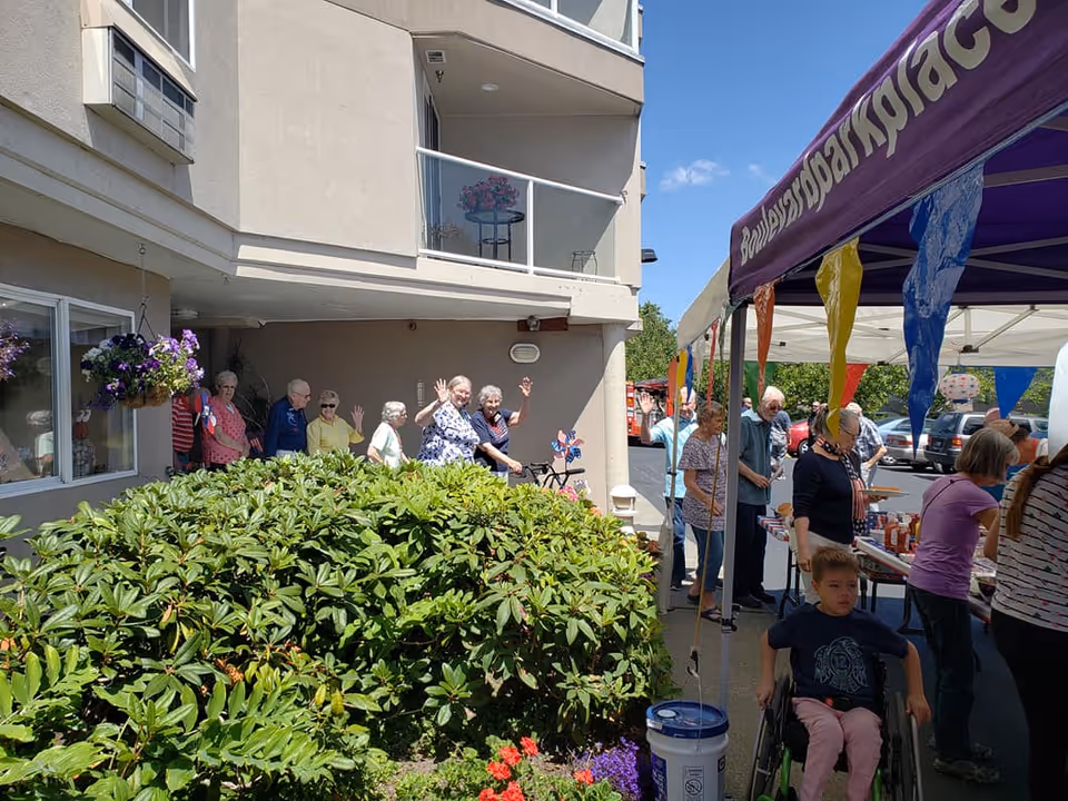 A group of elderly people and some younger individuals gathered outside a building under a tent with colorful pennant banners. Some elderly people are standing near the building, waving and smiling, while others are lined up at a table with food and drinks. A young child in a wheelchair is in the foreground. The building has balconies and hanging flower baskets.