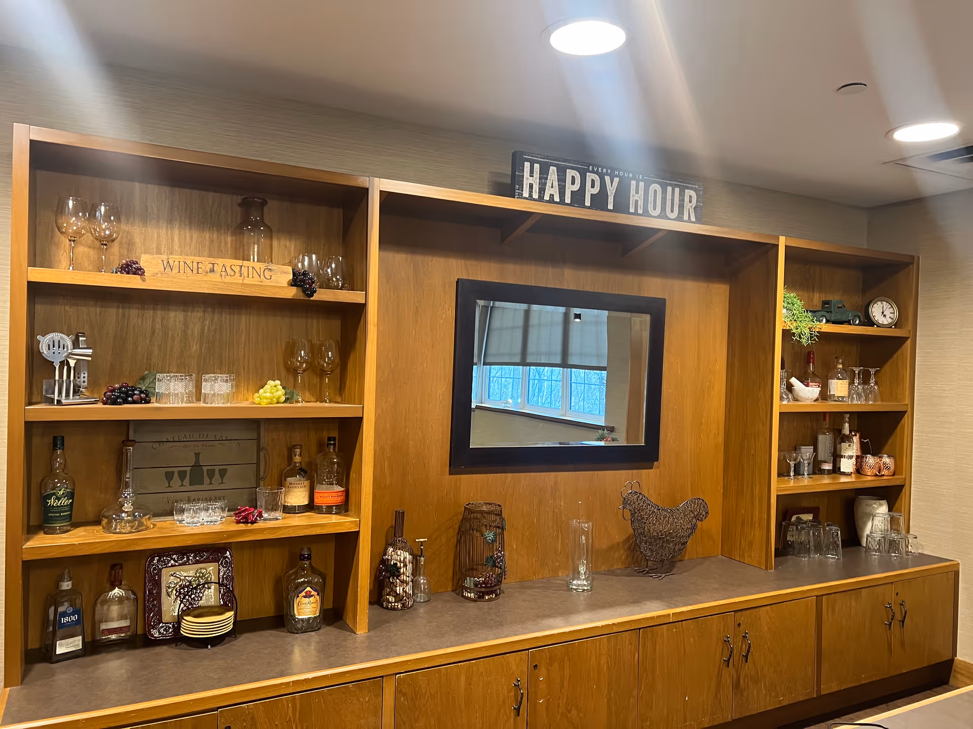 A wooden built-in bar area with shelves displaying various bottles of liquor, wine glasses, decorative grapes, and a sign that reads 'WINE TASTING'. Above the bar is a sign that says 'HAPPY HOUR'. There is a mirror in the center reflecting a window with a view outside. The countertop has decorative items including a wire chicken sculpture and glass containers.