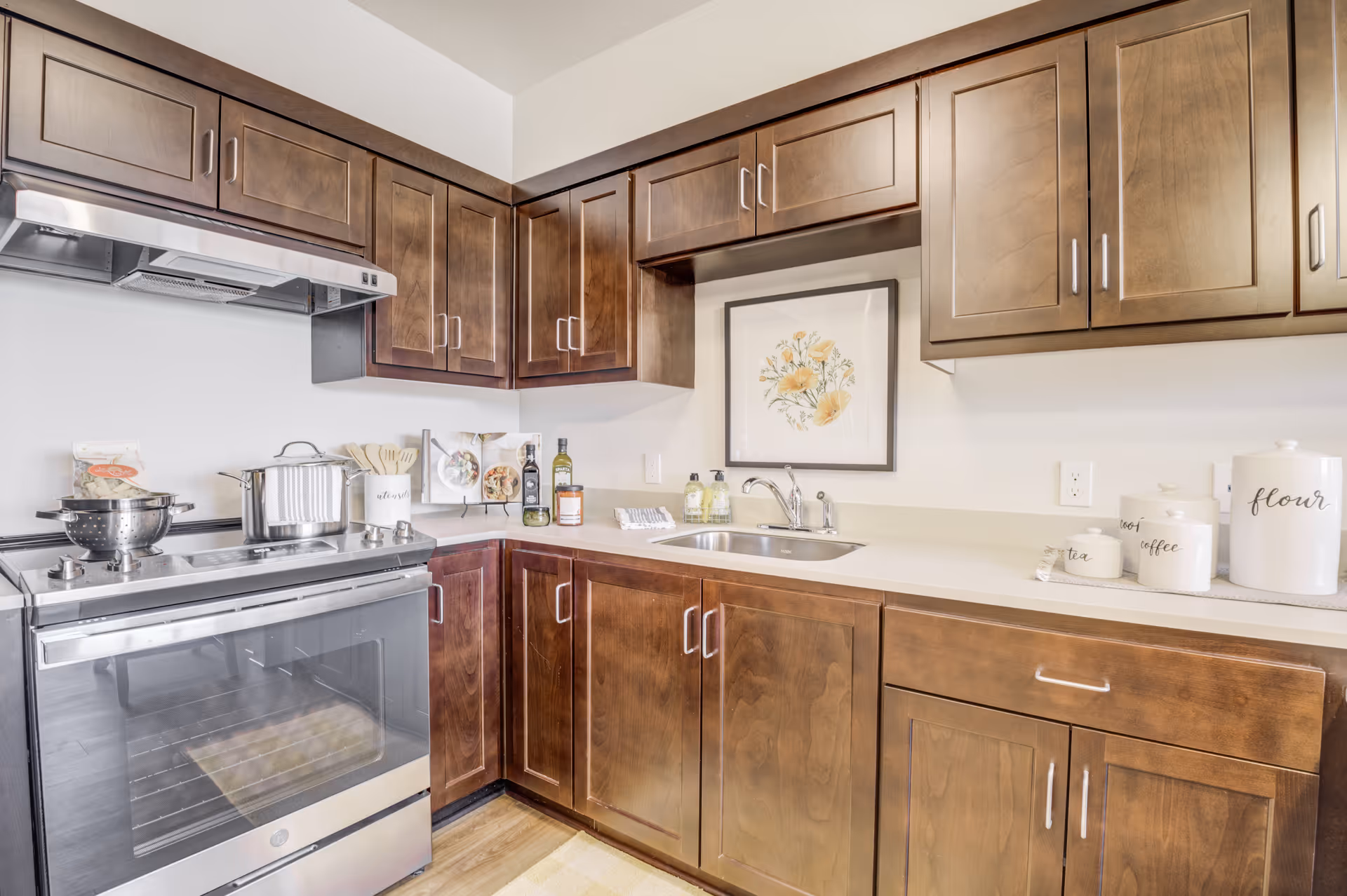 A modern kitchen with dark wooden cabinets, a stainless steel stove with pots on it, a countertop with a sink, decorative jars labeled tea, coffee, and flour, and a framed floral artwork on the wall.