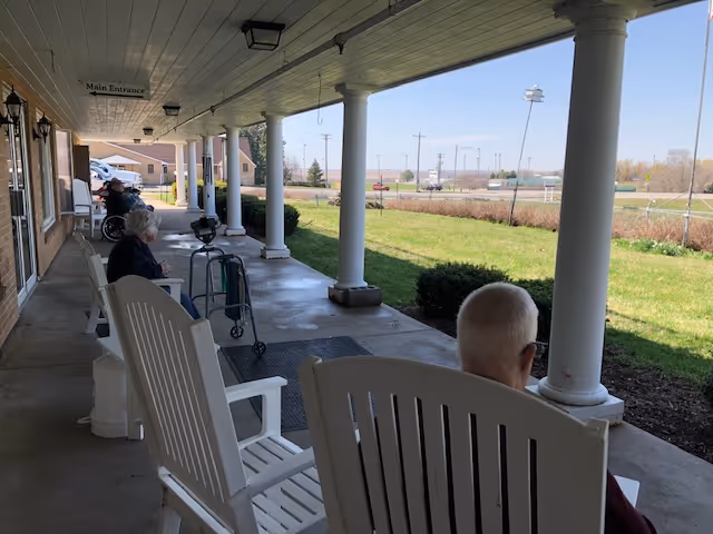 A covered outdoor patio area at Oakland Manor with white columns and several white rocking chairs. Two elderly individuals are seated in the chairs, facing a grassy area with bushes and a sports field in the background. The patio has a concrete floor and a ceiling with lights, and a sign indicating the main entrance is visible on the wall.