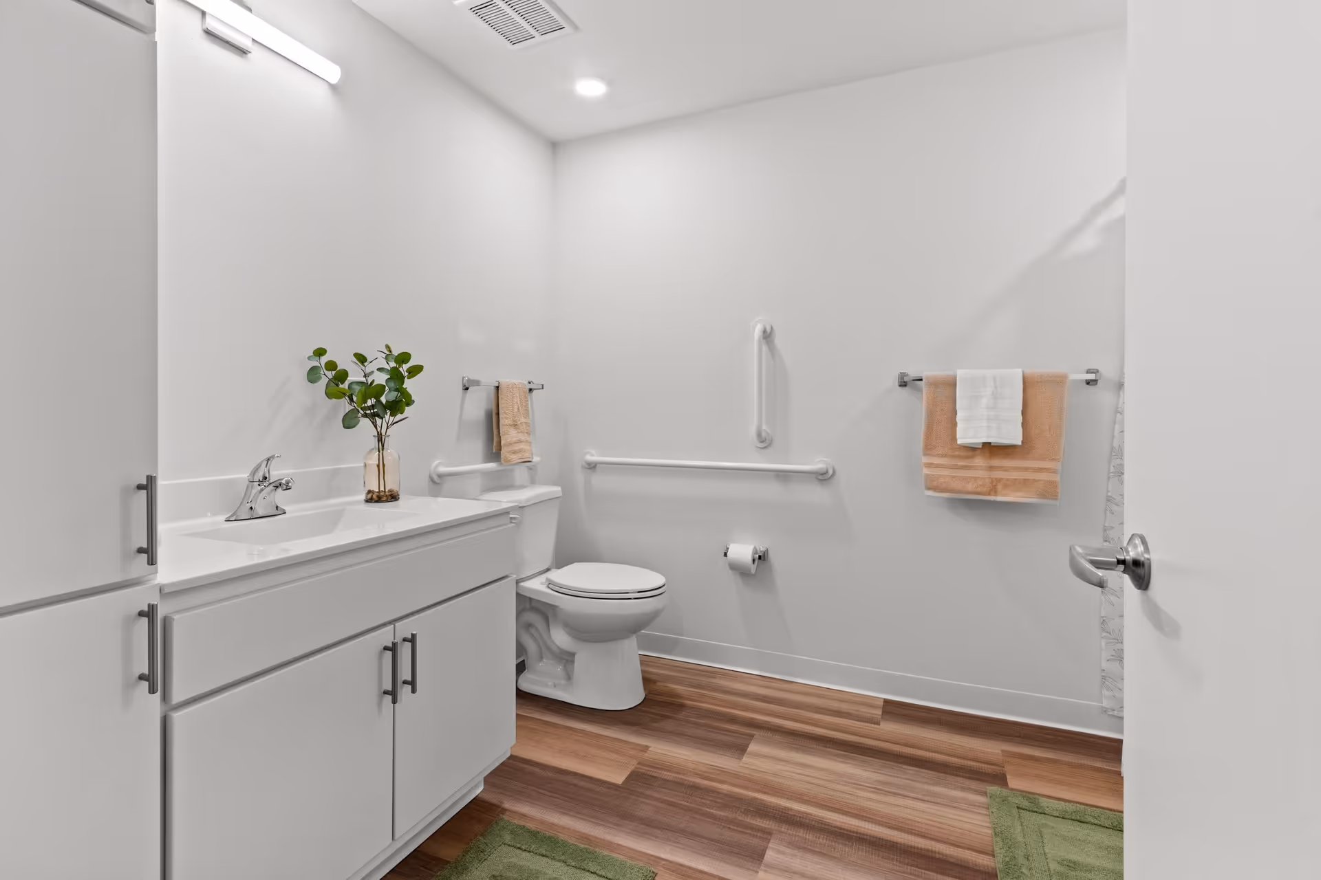 A clean, modern bathroom with white walls and wood-look flooring. There is a white vanity with a sink and a small vase with green leaves on the countertop. A toilet is positioned next to the vanity, with grab bars installed on the walls for accessibility. Two towel racks hold beige and white towels. The bathroom door is partially open, and there are green bath mats on the floor.
