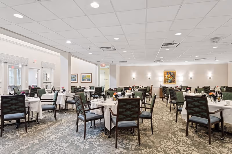A spacious dining room with multiple round tables covered with white tablecloths, each set with plates, cups, and floral centerpieces. The room has green cushioned chairs, patterned carpet, soft wall lighting, and framed artwork on the walls.