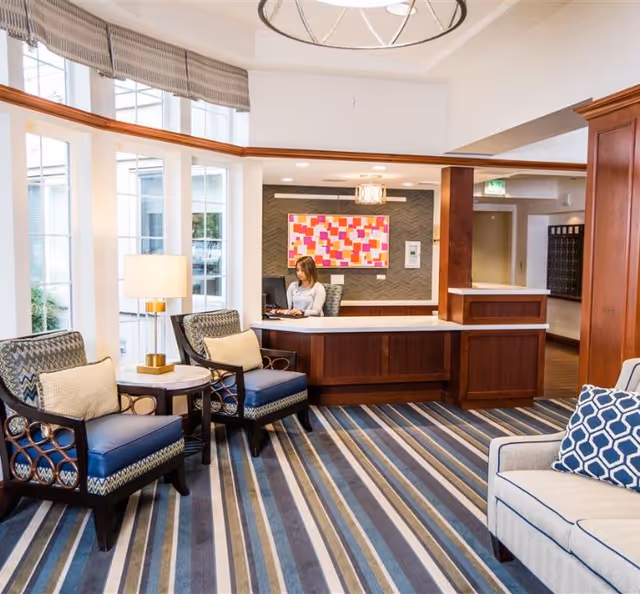 A bright and welcoming reception area with a woman seated behind a wooden front desk working on a computer. The room features large windows with striped valances, two patterned armchairs with cushions, a small round table with a lamp, and a beige sofa with a blue patterned pillow. The floor is covered with a striped carpet in shades of blue, gray, and beige. A colorful abstract painting hangs on the wall behind the desk.