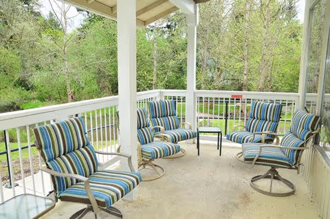 Covered outdoor patio area with five striped cushioned swivel chairs arranged around a small black table, overlooking a green wooded area.