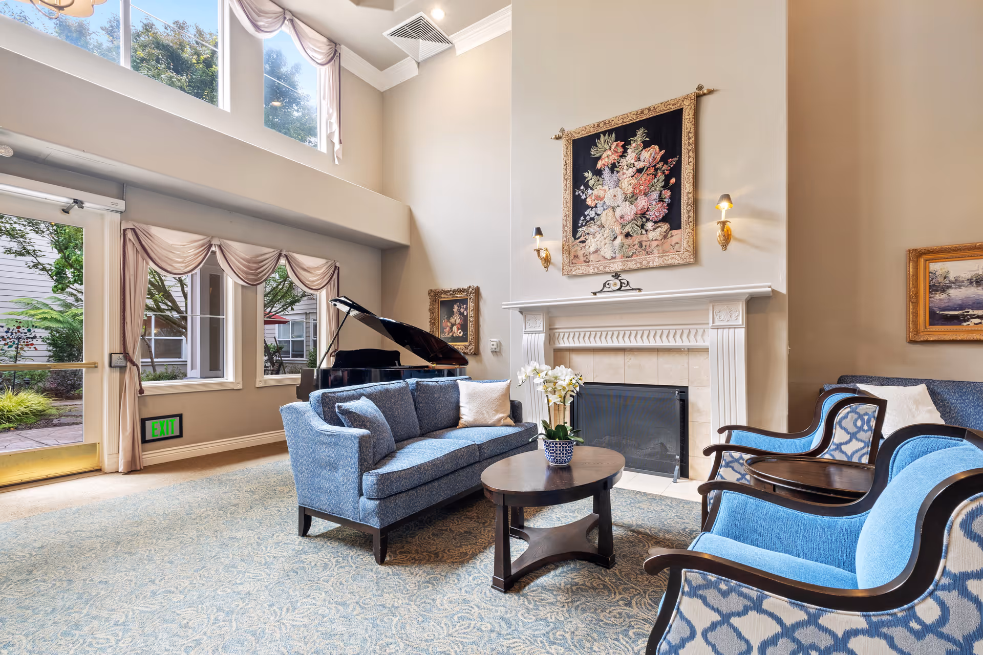 A bright and elegant living room with high ceilings and large windows. The room features a blue upholstered sofa and matching armchairs around a wooden coffee table with a potted orchid. A grand piano is positioned near the windows, and a decorative fireplace with floral artwork above it is on the wall. The room is decorated with framed paintings and has soft beige walls and a patterned carpet.
