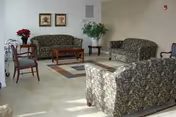 A senior living facility common area with floral patterned sofas and chairs arranged around two wooden coffee tables on a tiled floor. There are two framed pictures on the wall and a large potted plant in the corner.