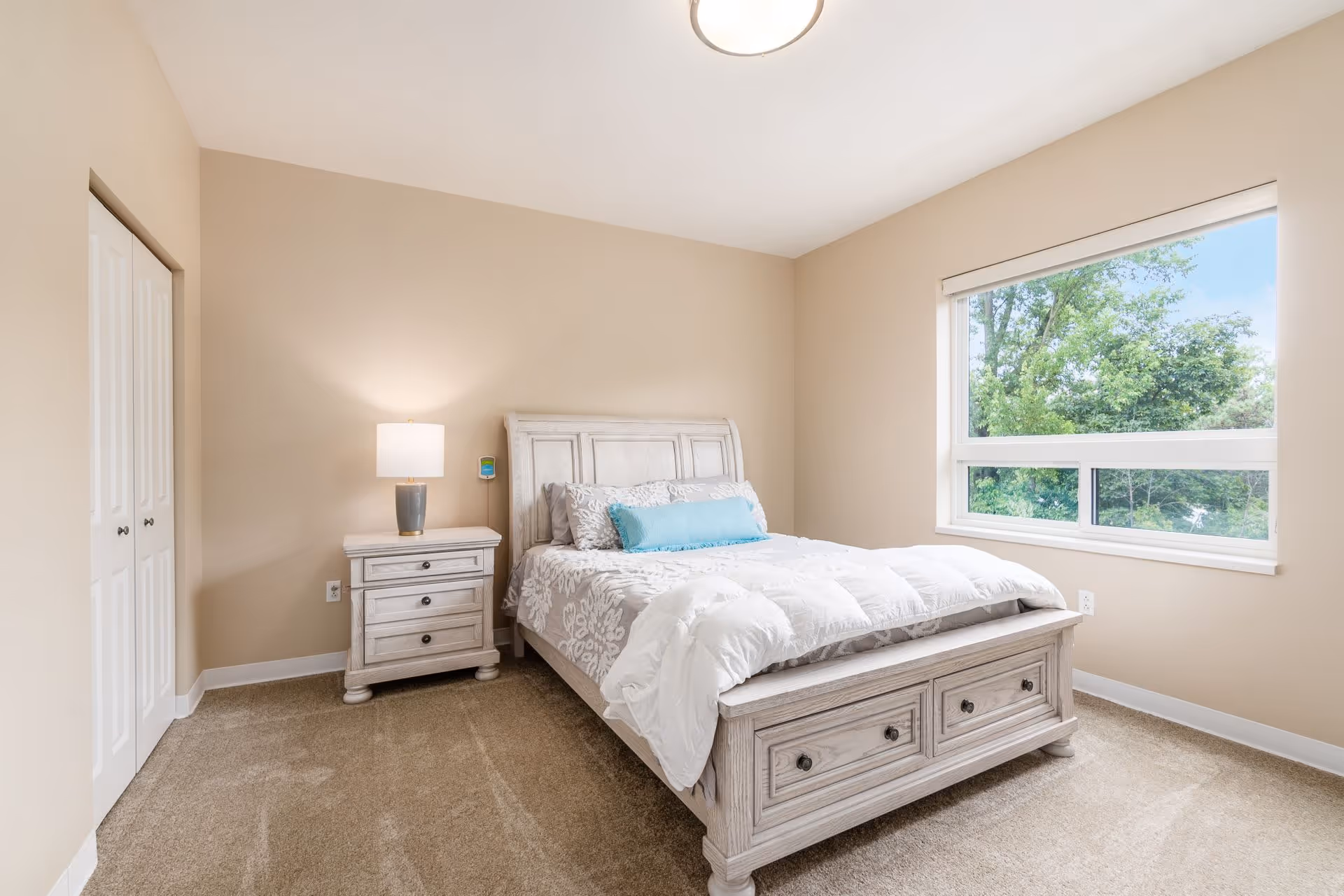 A bright bedroom with beige walls and carpeted floor featuring a white wooden bed with a white and gray patterned comforter and a blue accent pillow. Next to the bed is a matching white wooden nightstand with a lamp on it. A large window shows green trees outside, and there is a closed white closet door on the left wall.