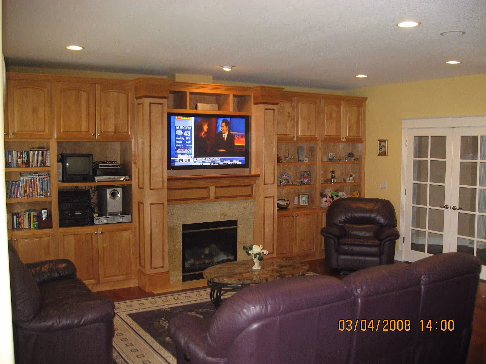 A cozy living room with wooden built-in cabinets and shelves surrounding a fireplace. A flat-screen TV is mounted above the fireplace, showing a news program. The room has leather seating including a sofa and armchair, a coffee table with a small flower arrangement, and French doors on the right side. The walls are painted yellow and the floor has a patterned area rug.
