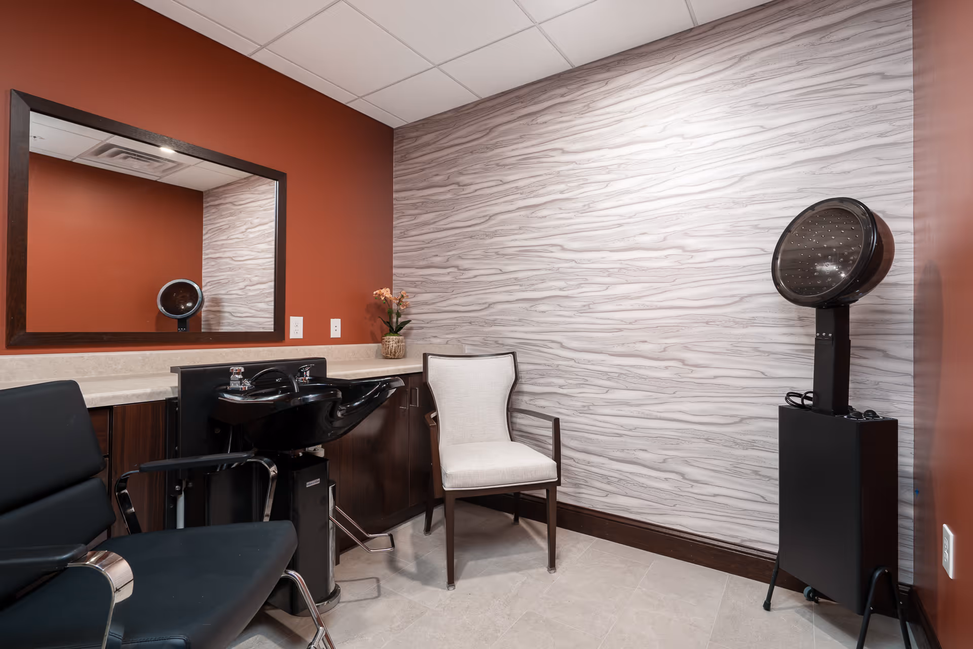 Salon-style room with a hair-wash basin, styling chairs, a hooded dryer, a mirror, and an upholstered chair against a wood-patterned wall.
