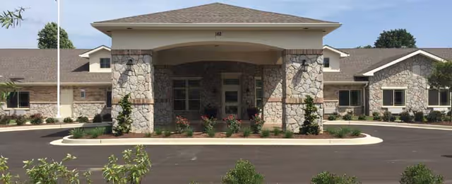 Front exterior view of Hickory Hills Alzheimer's Special Care Center featuring a stone facade building with a covered entrance supported by stone pillars, surrounded by a paved driveway and landscaped greenery.