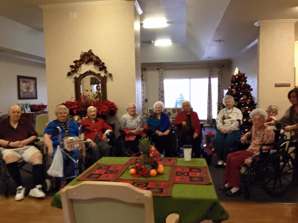 A group of elderly people sitting in a common area of an assisted living facility. They are seated in a semi-circle around a table decorated with a green tablecloth, a centerpiece of fruit, and a red holiday-themed runner. There is a Christmas tree with decorations in the background, along with poinsettia plants and a mirror on the wall. The room has a warm and cozy atmosphere with natural light coming through a large window.