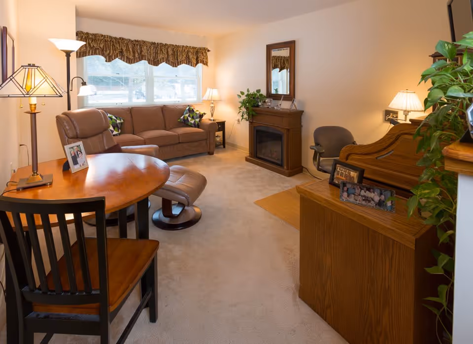 A cozy living room with a brown sofa, a reclining chair, a wooden table with a chair, a floor lamp, a small electric fireplace with a mirror above it, and various framed photos and plants. The room has beige walls and carpet, with a window covered by a patterned valance.