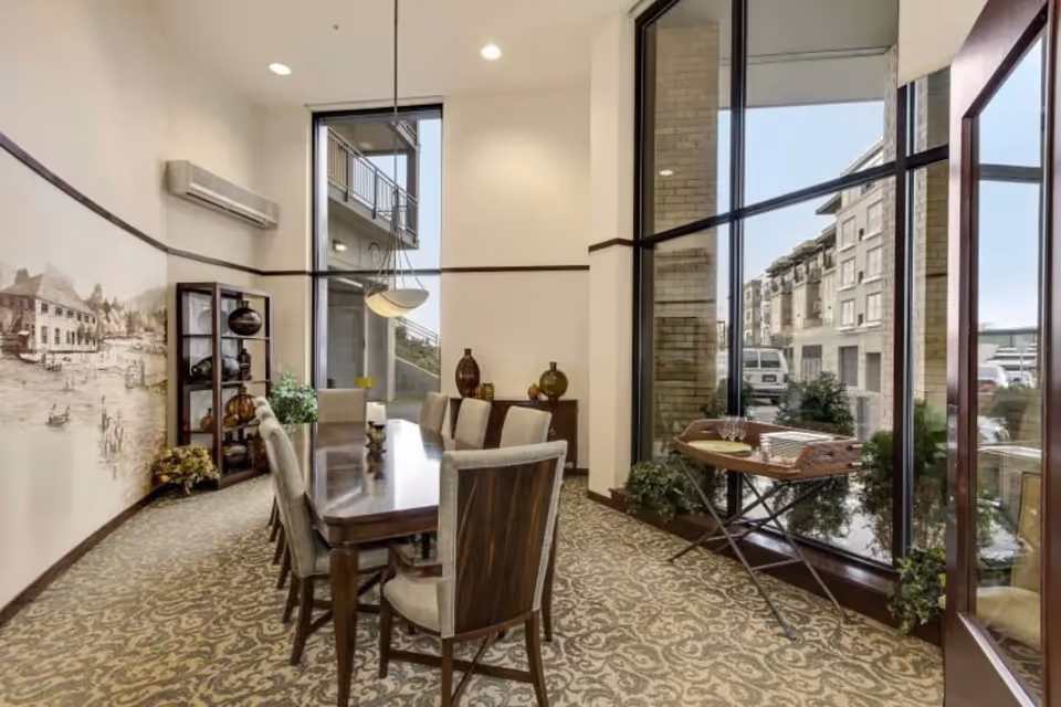 A well-lit dining room with a long wooden table surrounded by eight upholstered chairs. The room features large floor-to-ceiling windows on one side, allowing natural light to fill the space and offering a view of nearby buildings. There is a decorative shelving unit with vases and plants against one wall, and a mural depicting a village scene. The carpet has an intricate pattern, and a small table near the windows holds stacked plates and glassware.