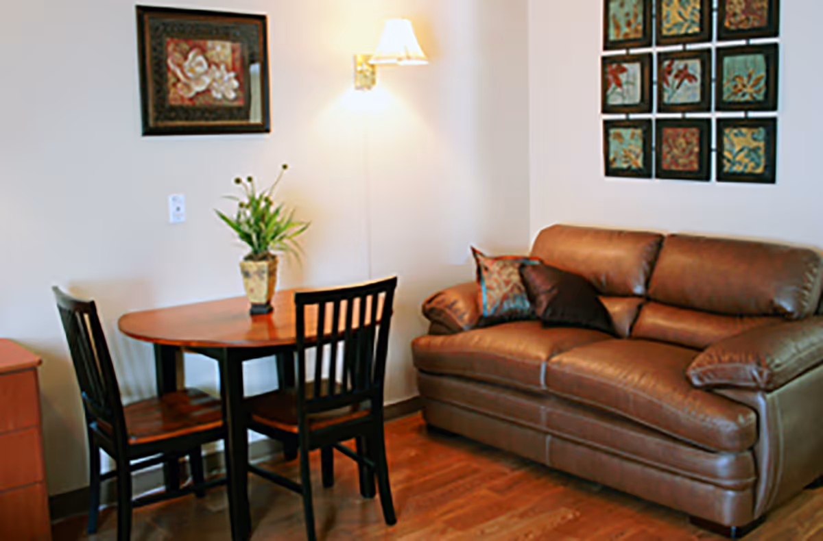 Small living area with a brown leather loveseat, a round wooden table with two chairs and wall art.