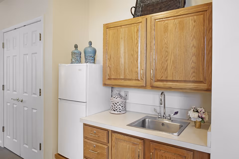 Small kitchen area with a white refrigerator, wooden cabinets, a countertop with a stainless steel sink, and decorative items.