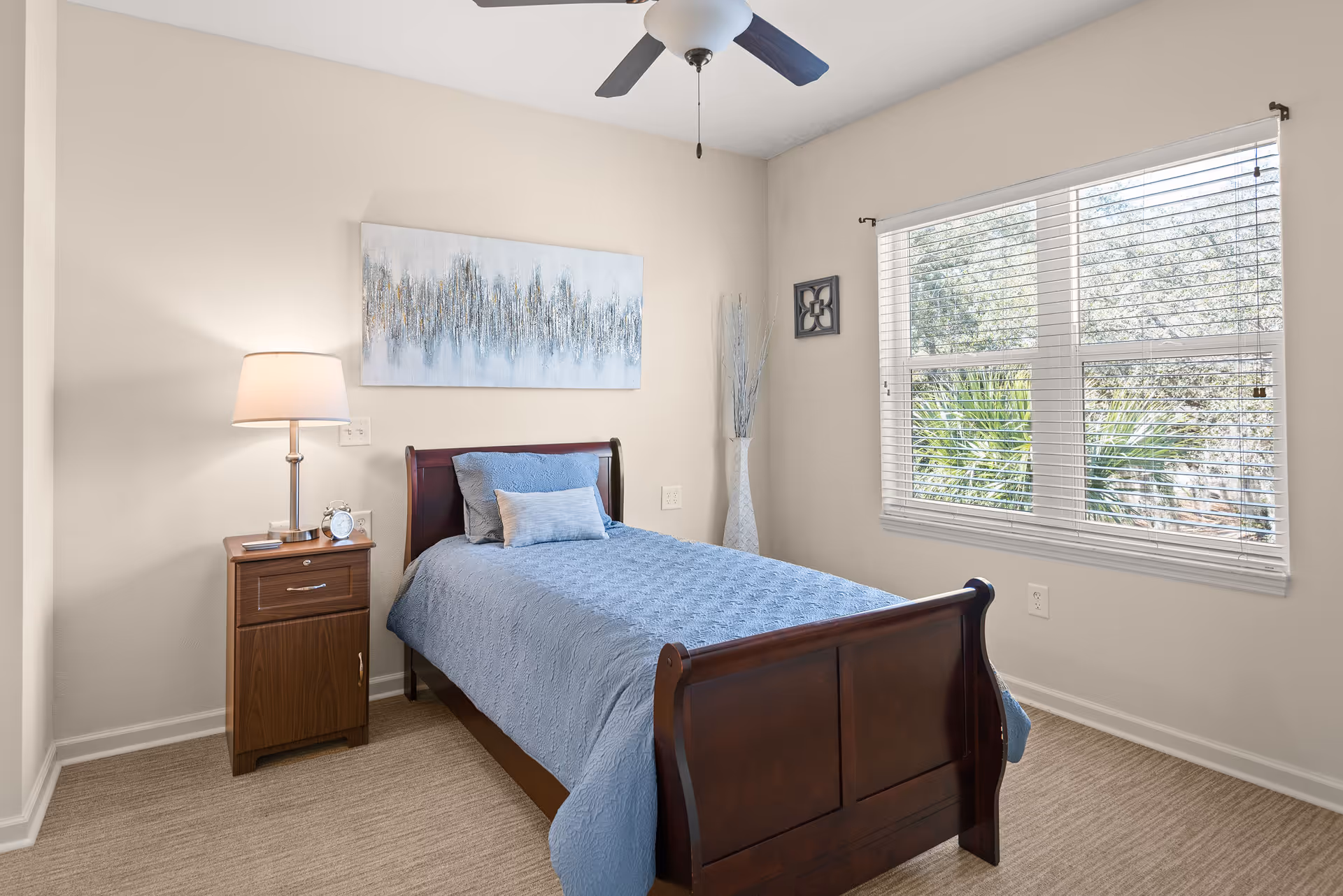 Sunlit small bedroom with a single wooden sleigh bed dressed in blue bedding, a nightstand with a lamp and clock, wall art, and a window with blinds.