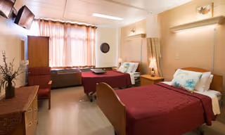 A well-lit room with two single beds covered in red bedspreads, each with white pillows and decorative cushions. Between the beds is a wooden nightstand with a lamp. A window with sheer curtains lets in natural light. The room also features a wooden dresser with a vase of flowers, a wall-mounted TV, and a chair.
