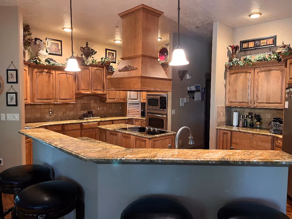 Open kitchen with wooden cabinets, a granite-topped island and breakfast bar with three black stools, pendant lights, and a stainless sink.