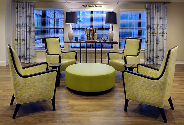 A lounge area with four yellow armchairs arranged around a round green ottoman on a wooden floor. Behind the chairs is a console table with two matching lamps and decorative items, positioned in front of three large windows with white blinds and patterned curtains. A sign above the windows reads 'Philip Launer Lounge'.