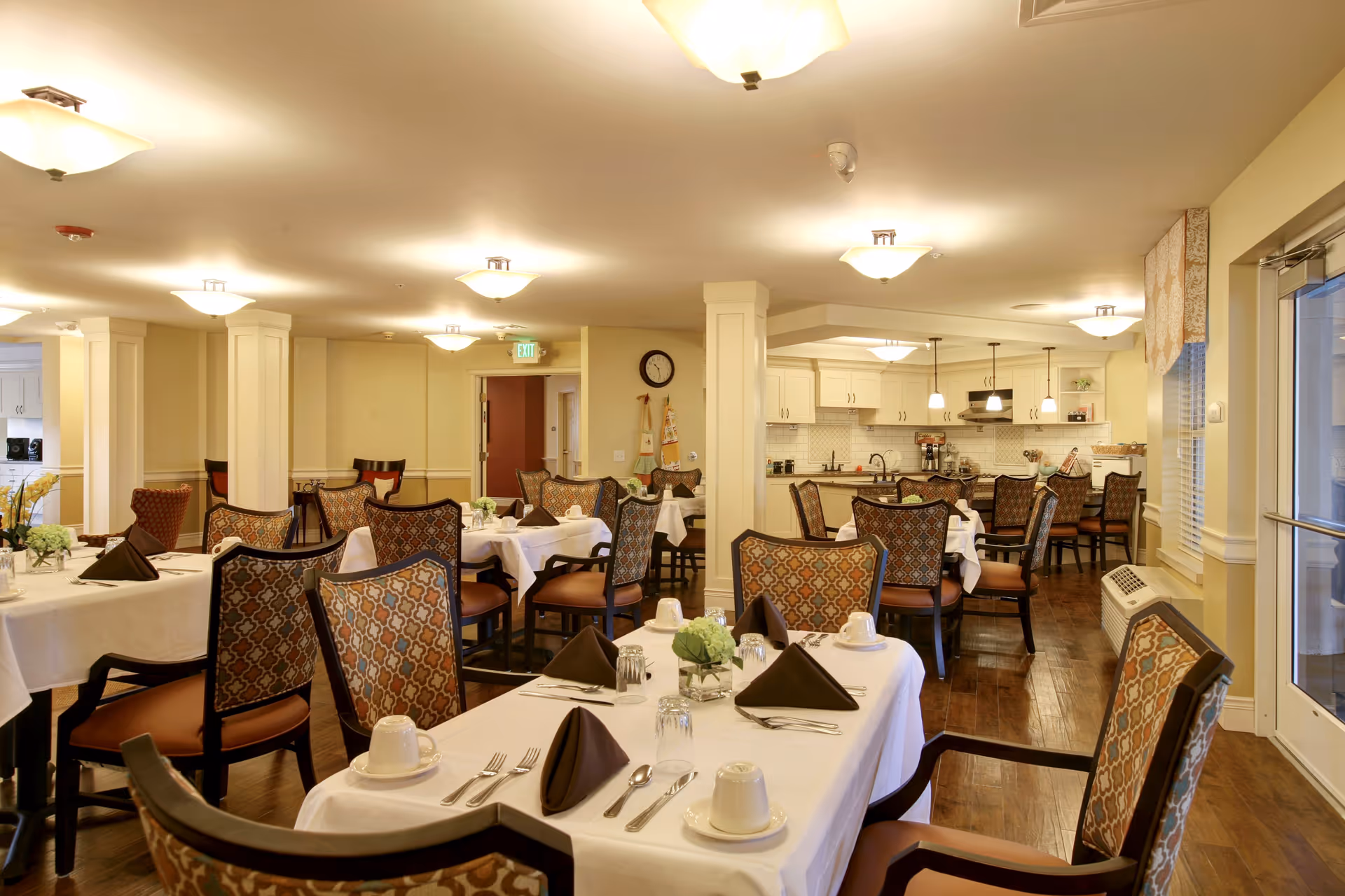 Warmly lit dining room with multiple tables set with white linens, napkins, and place settings and upholstered chairs, with a kitchen area visible in the background.