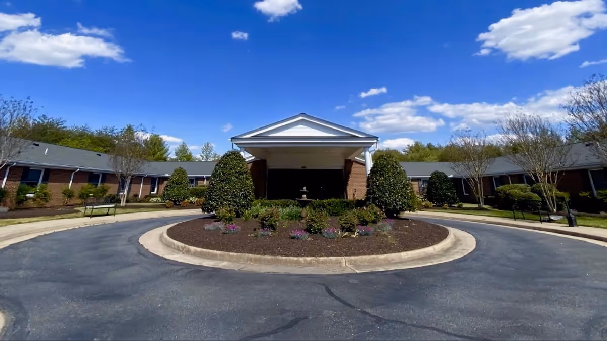 Front exterior view of a single-story assisted living facility with a circular driveway and landscaped garden in the center. The building has a covered entrance with white columns and a triangular pediment. The sky is blue with scattered clouds.