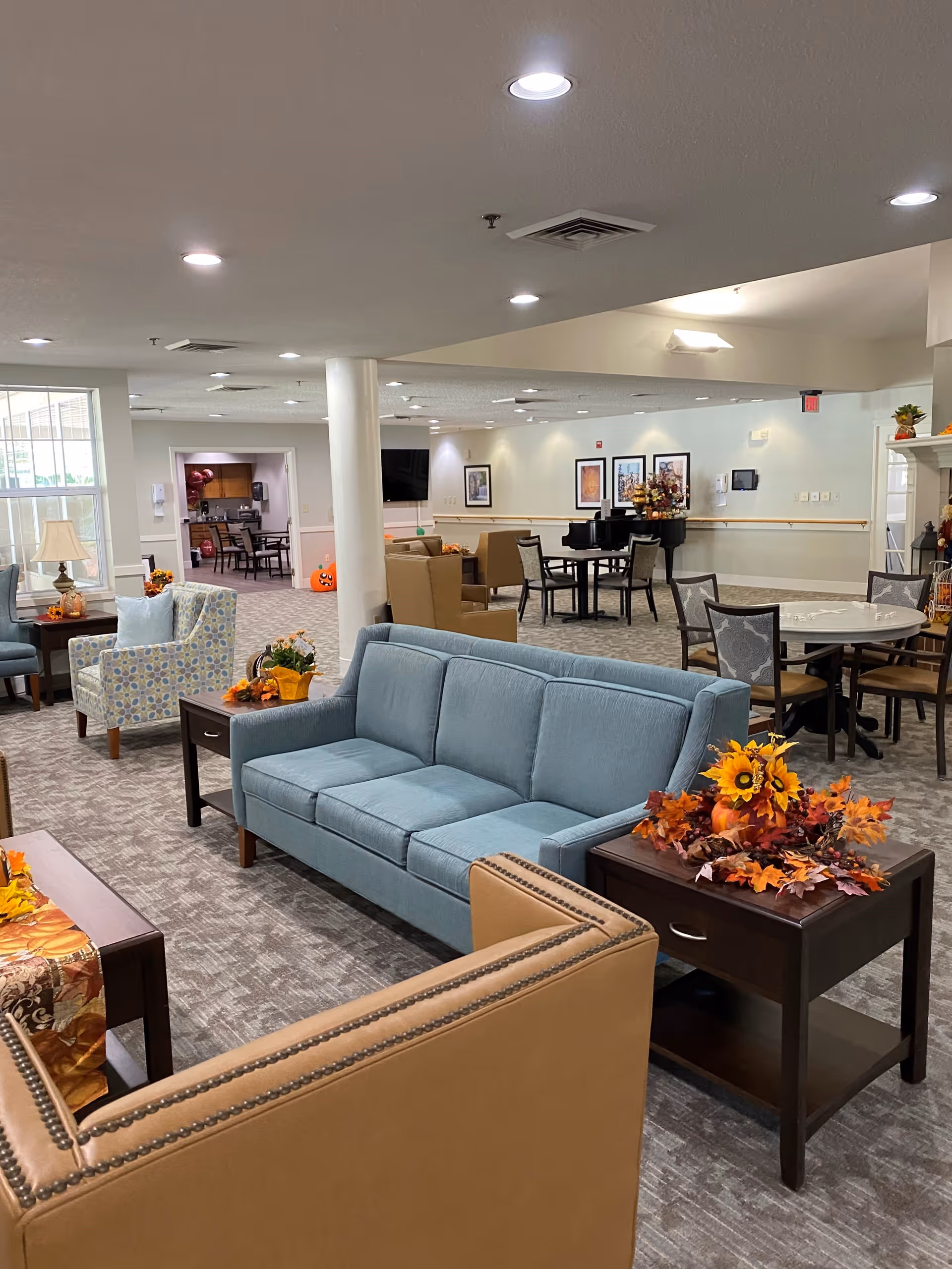 A spacious senior living facility common area with various seating options including a blue sofa, patterned armchairs, and tan chairs. There are side tables decorated with autumn-themed floral arrangements. In the background, there are more tables and chairs, a piano, framed artwork on the walls, and a doorway leading to another room. The space is well-lit with ceiling lights and has a carpeted floor.